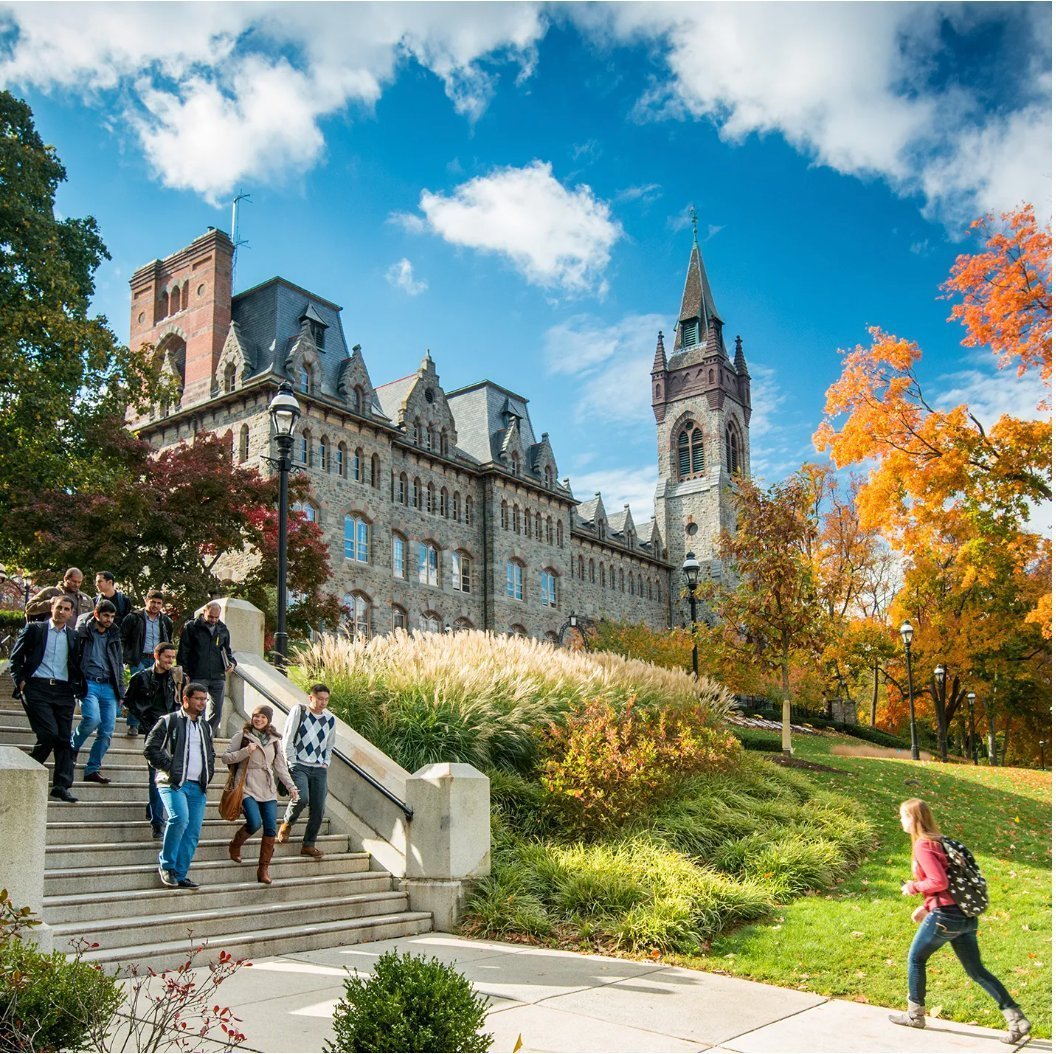 University campus in autumn — students walking down stone steps from a Gothic Revival hall with a stone tower and golden foliage
